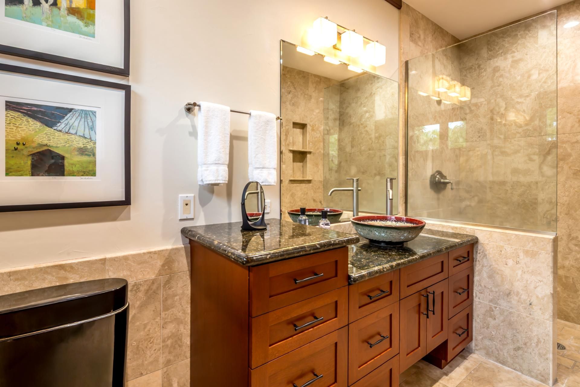 A bathroom with a marble vanity, a large mirror, a glass-enclosed shower, and framed artwork on the wall.