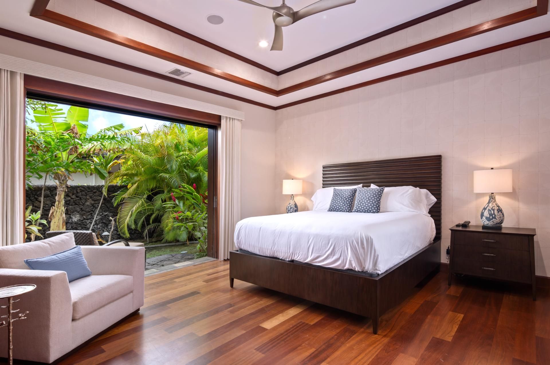 A bedroom with a large bed, a nightstand, a lamp, a chair, and a view of tropical plants through a glass door.