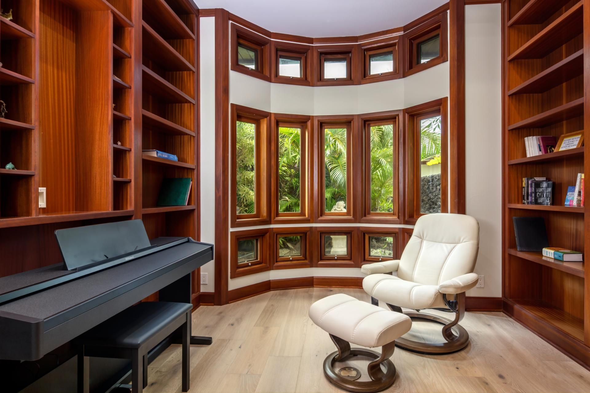 A library with wooden bookshelves, a piano, a reclining chair, and large windows with a view of greenery.