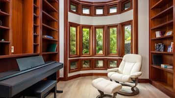A library with wooden bookshelves, a piano, a reclining chair, and large windows with a view of greenery.