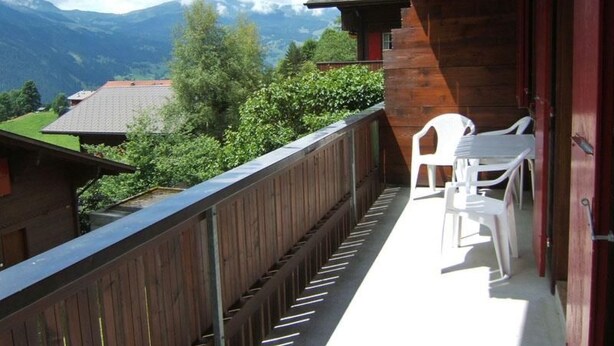 A balcony with a wooden railing, white plastic chairs, and a view of mountains and greenery. - Chrinnenhorn 4 Bett Wohnung by Interhome (Grindelwald)