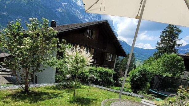 A patio with a metal table and chairs, a white umbrella, and a view of a mountain and a house in the background. - Apartment Aelpli by Interhome (Grindelwald)