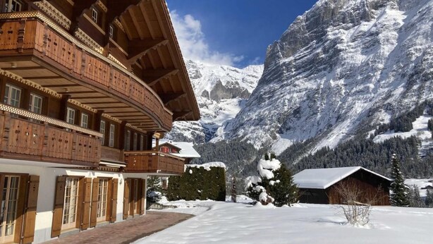 Een klassiek houten chalet met een gebogen balkon, tegen een achtergrond van met sneeuw bedekte bergen.