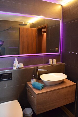 Un bany modern amb un moble de tocador de fusta, un lavabo blanc i un mirall gran amb il·luminació LED morada.