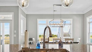 A kitchen with granite countertops, stainless steel sink, pendant lights, and a view through large windows. - Isle Have Another by Brunswick Vacation Rentals: 7BR w/ Elevator, Pool & Views! (Ocean Isle Beach)