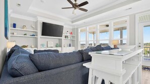 A living room with a grey sofa, a white table, a ceiling fan, and a TV. - Isle Be Back by Brunswick Vacation Rentals: Oceanfront, Pool, Elevator & Views! (Ocean Isle Beach)
