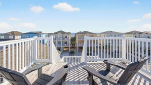 A wooden deck with outdoor seating, a view of a residential area with multi-story buildings, and a clear sky. - Isle Be Back by Brunswick Vacation Rentals: Oceanfront, Pool, Elevator & Views! (Ocean Isle Beach)