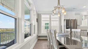 A modern kitchen with large windows, a dining area with white chairs, and stainless steel appliances. - Isle Be Back by Brunswick Vacation Rentals: Oceanfront, Pool, Elevator & Views! (Ocean Isle Beach)