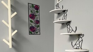 Un coin de pièce avec un mur blanc, une œuvre d’art florale, un pot de plante blanc et une étagère blanche fixée au mur, ornée de motifs géométriques.