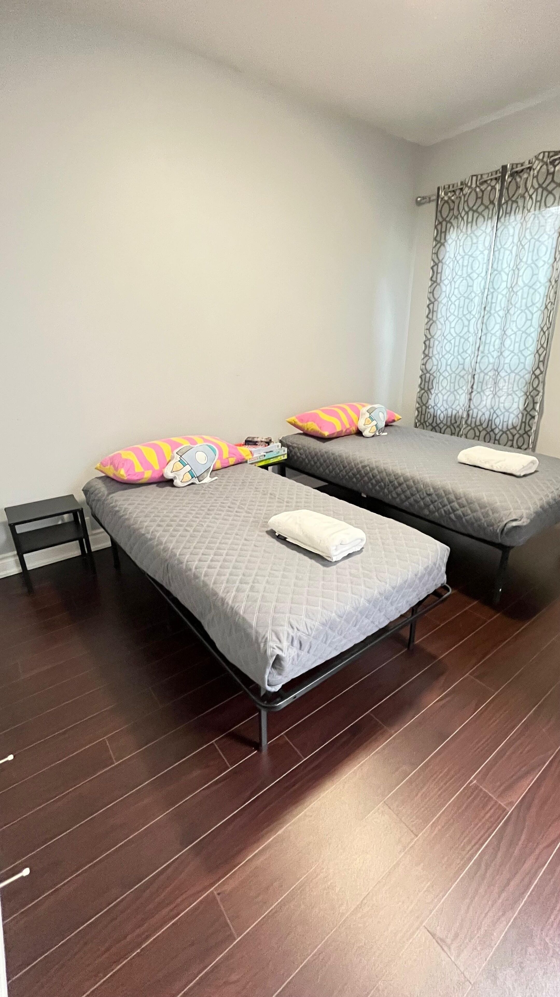 Ein Zimmer mit zwei Einzelbetten, jedes mit einer grauen Steude und einem weißen Kopfkissen. Neben den Betten steht ein kleiner Nachttisch, und ein Fenster mit gemusterten Vorhängen.
