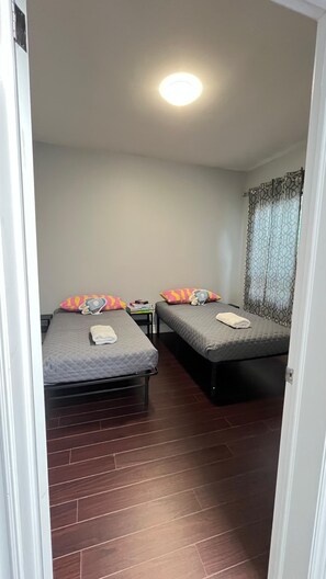 A small, well-lit room with two beds, each with a pillow and a small table. The room has a wooden floor and a plain wall with a window covered by patterned curtains. - Cozy Apartment in the Heart of Kensington Market (Toronto)