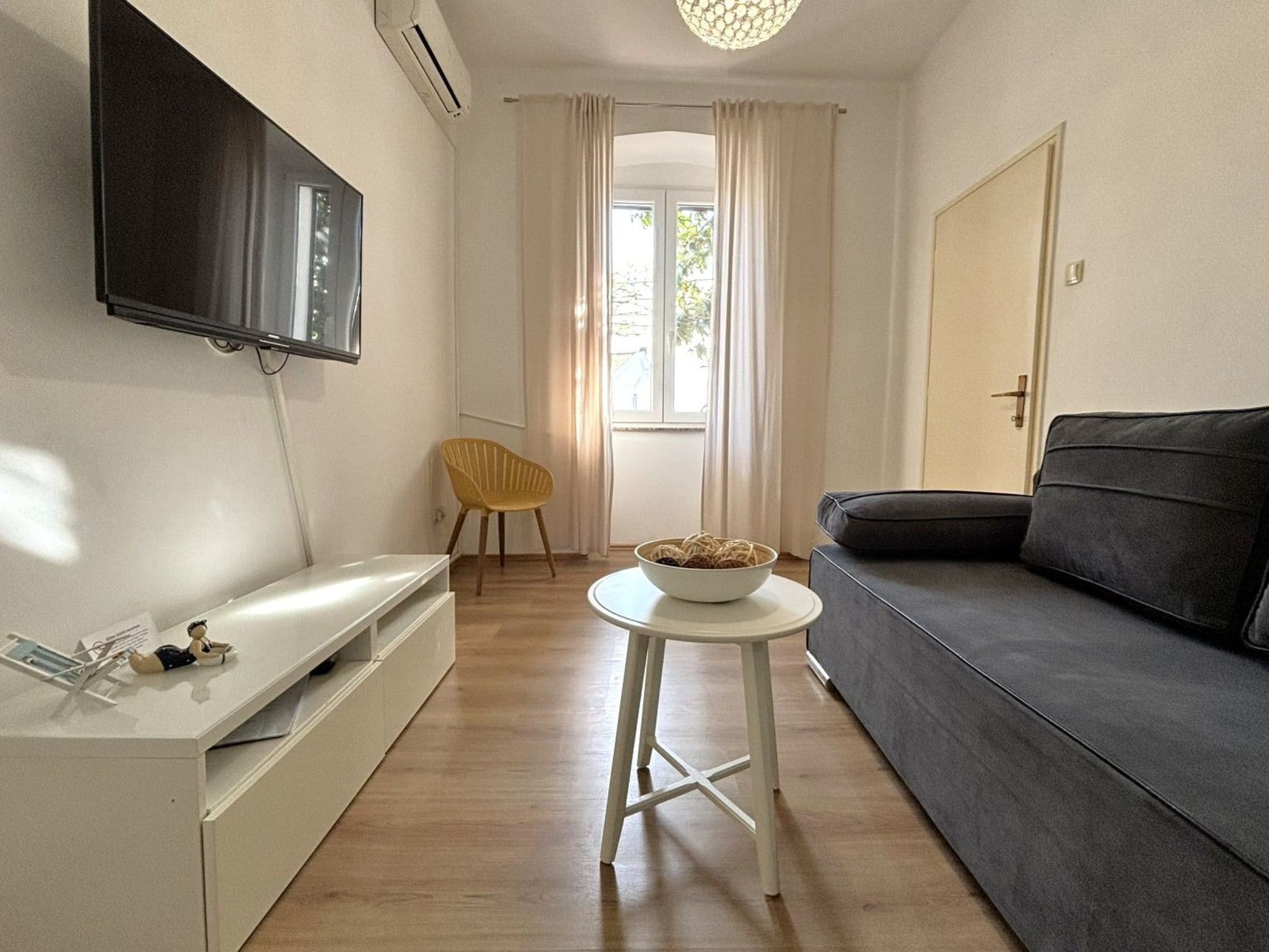 Un espace de vie compact comprenant un téléviseur à écran plat fixé au mur, un canapé, une petite table ronde avec un saladier, une chaise jaune et un meuble blanc.