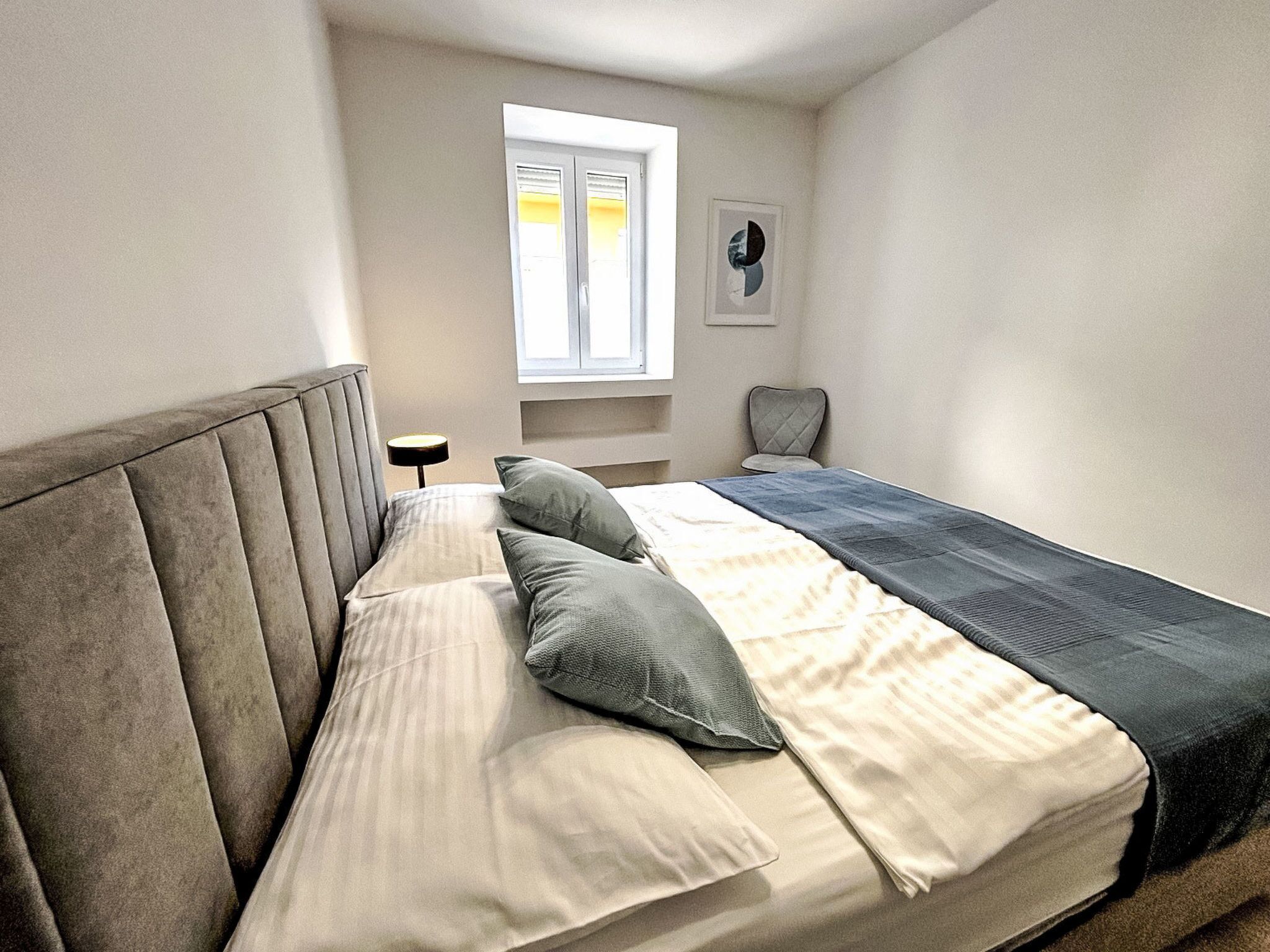 Een slaapkamer met een bed, kussens, een stoel, een nachtkastje, een raam en een wandplank.