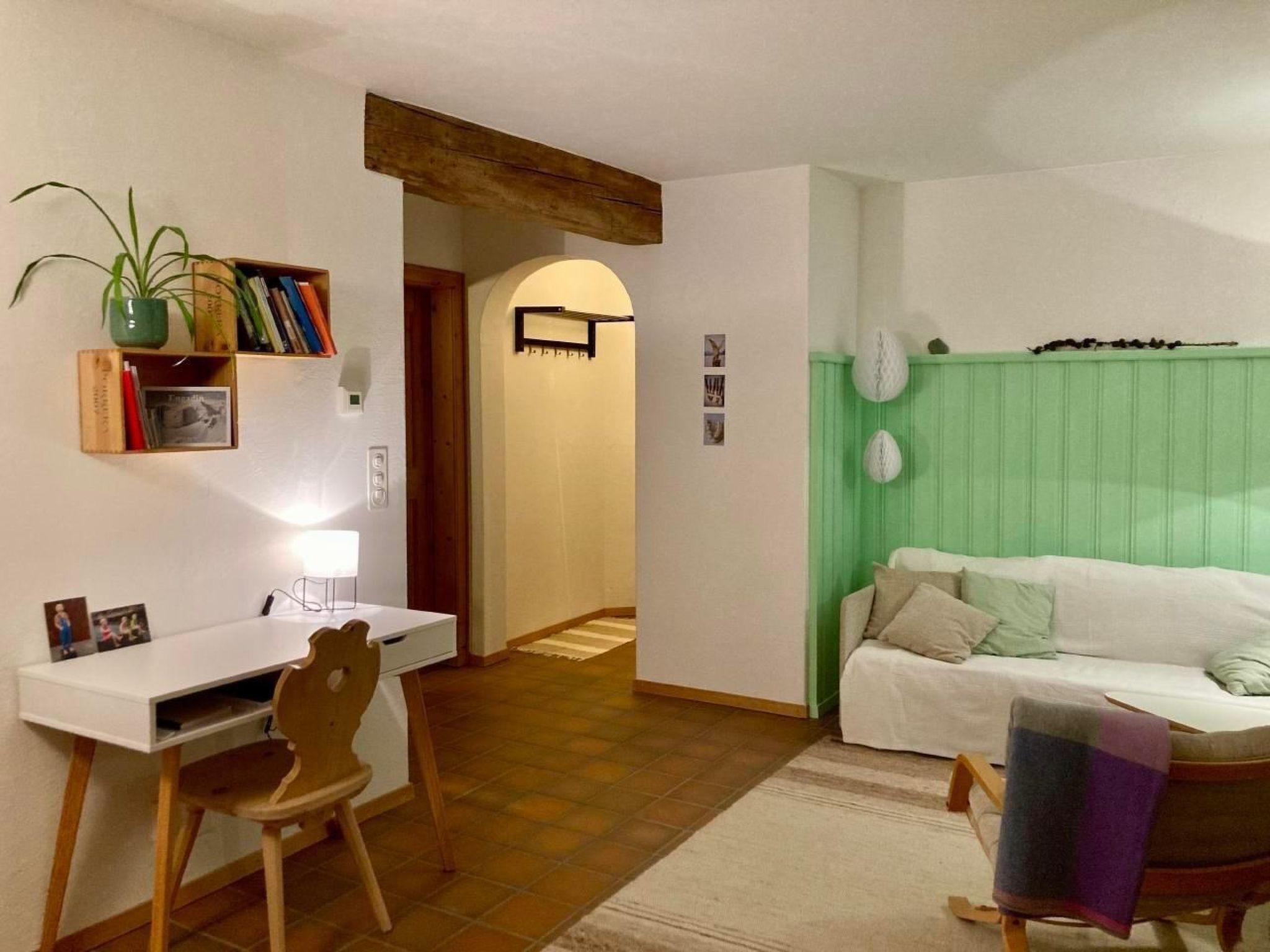 Ein gemütliches Wohnzimmer mit einem weißen Sofa, einem Schreibtisch mit Stuhl, einer grünen Akzentwand und einer Treppe, die in einen anderen Raum führt.