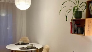 En matplats med ett runt bord, två stolar, en vit vägg, en bokhylla med böcker, en krukväxt och en hängande lampa.