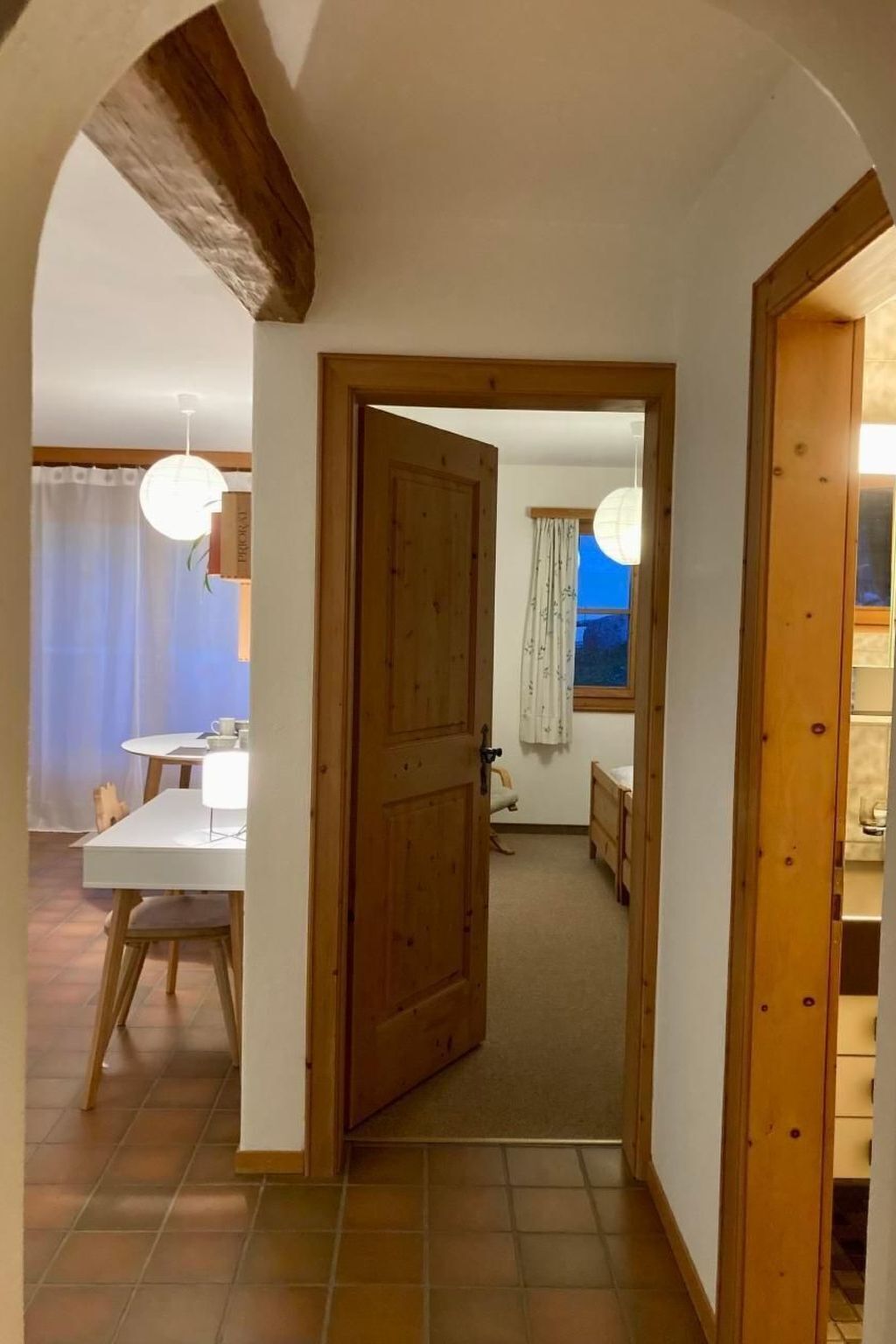 A hallway leading to a bedroom with a wooden door, a dining area with a table and chairs, and a window with a view.