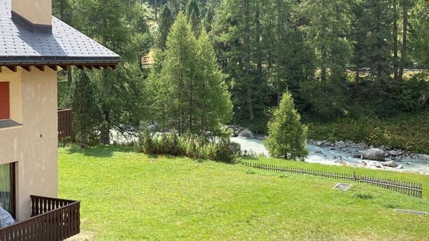 Una casa con un balcone che si affaccia su un prato e un fiume, circondata da una fitta foresta.