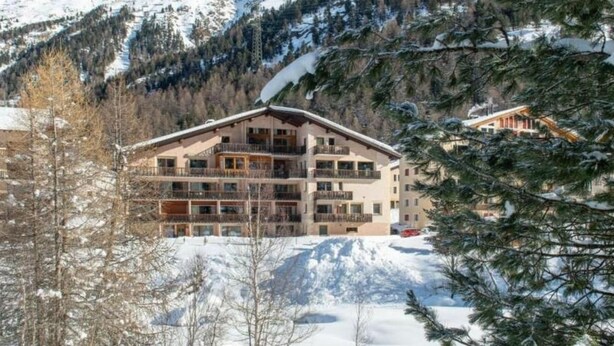 Un hôtel à plusieurs étages, doté de balcons, qui se dresse sur un fond de montagnes enneigées et de pins.