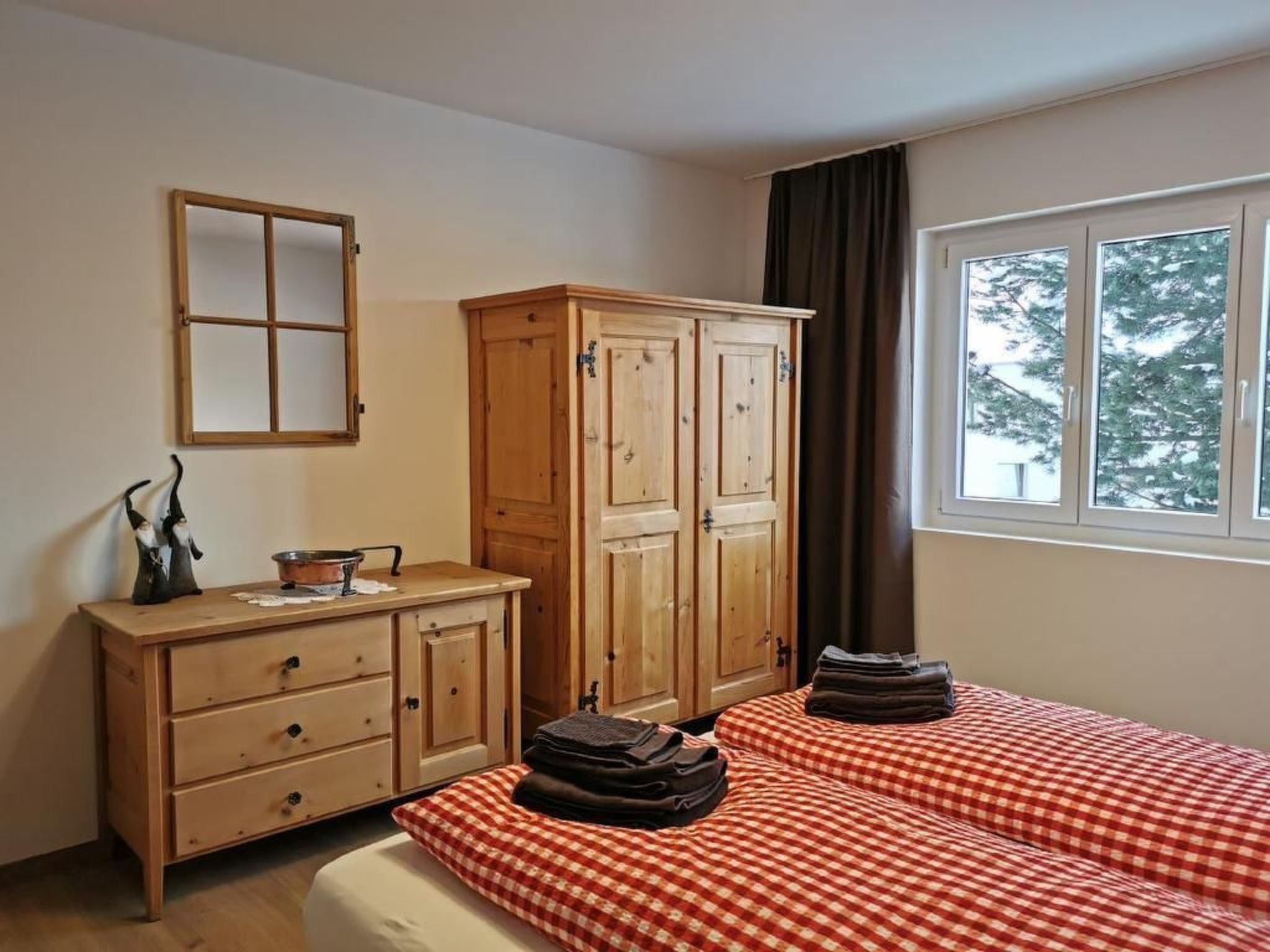 A bedroom with a wooden wardrobe, a bedside table, a mirror, and a window with a view of trees.