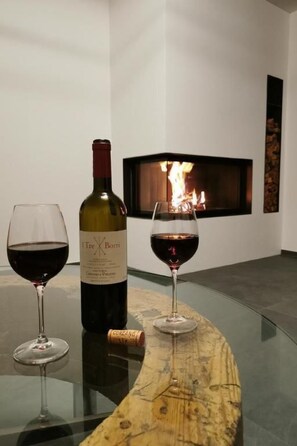 A bottle of wine and two glasses on a table with a wooden board, a fireplace in the background, and a stone wall. - Chesa Andrea, EG by Interhome (Pontresina)