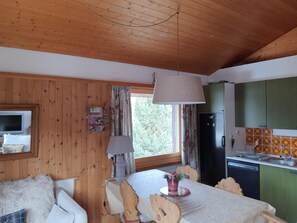 Een gezellig chaletinterieur met houten wanden, een eettafel met stoelen, een televisie en een keuken met groene kastjes en een zwarte koelkast.