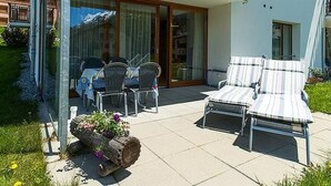 Un espace extérieur aménagé avec deux fauteuils et une table, un jardinière en bois remplie de fleurs et une porte vitrée donnant sur un balcon.