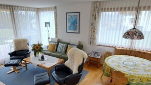 Un salon comprenant un canapé, deux fauteuils, une table basse et une table à manger recouverte d’une nappe à motifs floraux.