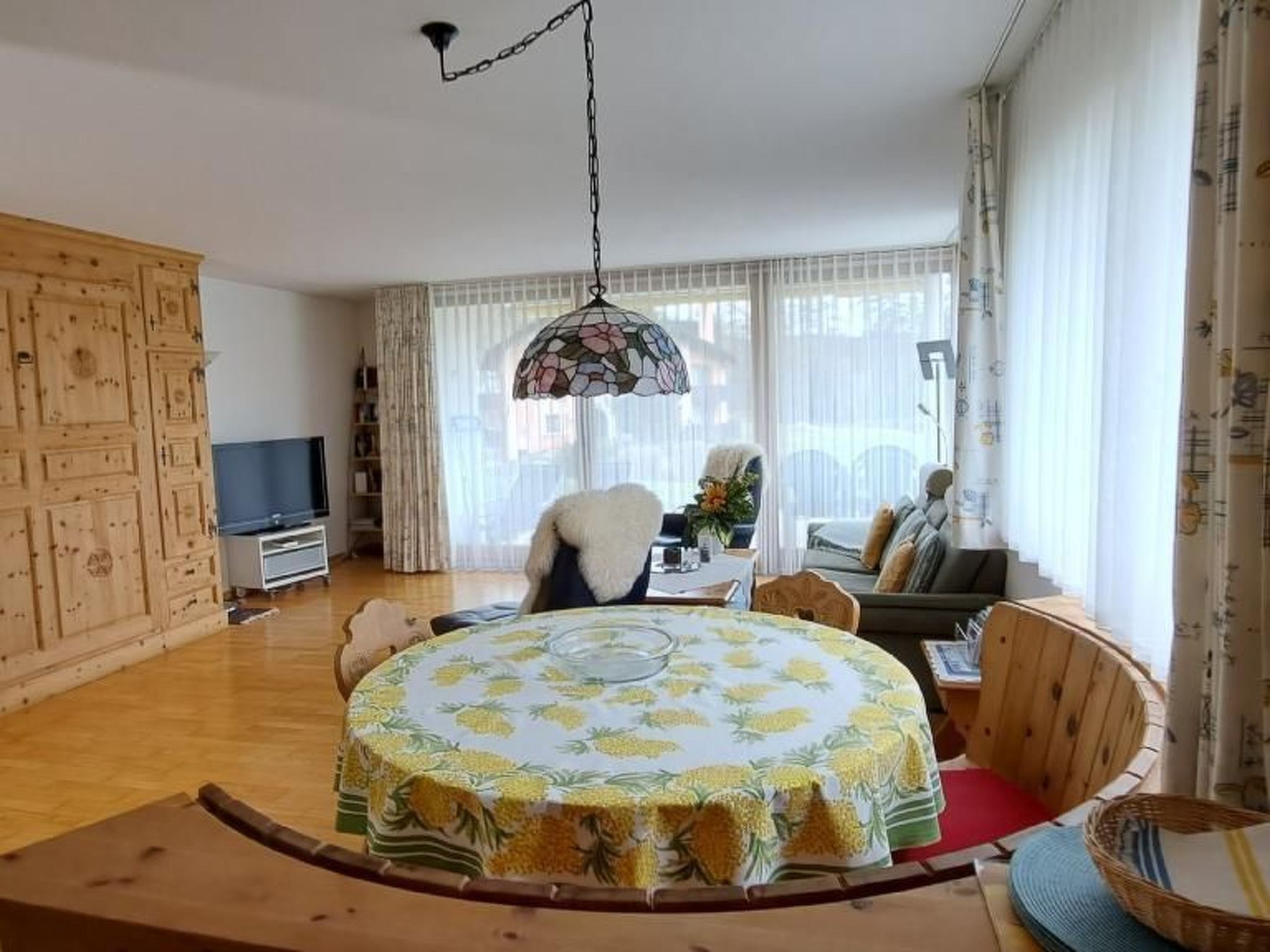Una sala de estar con una mesa redonda de madera cubierta con un mantel floral, un sofá, un televisor y un ventanal con cortinas.