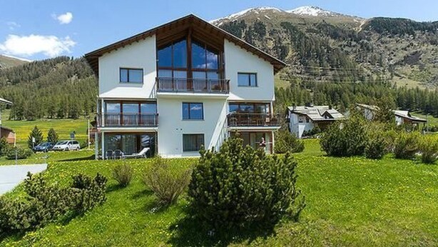 Un chalet à deux étages doté d’un balcon, entouré d’un paysage verdoyant et luxuriant, avec des montagnes en toile de fond.