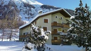 Un edifici de diverses plantes amb balcons, envoltat d'arbres i muntanyes cobertes de neu.