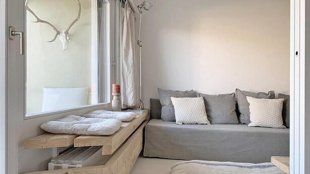 Un dormitori modern amb una panca integrada, un sofà gris amb molts coixins i una finestra gran amb cortines.