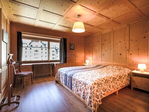 A wooden-paneled room with a bed, a desk, a chair, a window with a view of buildings, and a radiator. - Chesa Kober - adults only by Interhome (Pontresina)