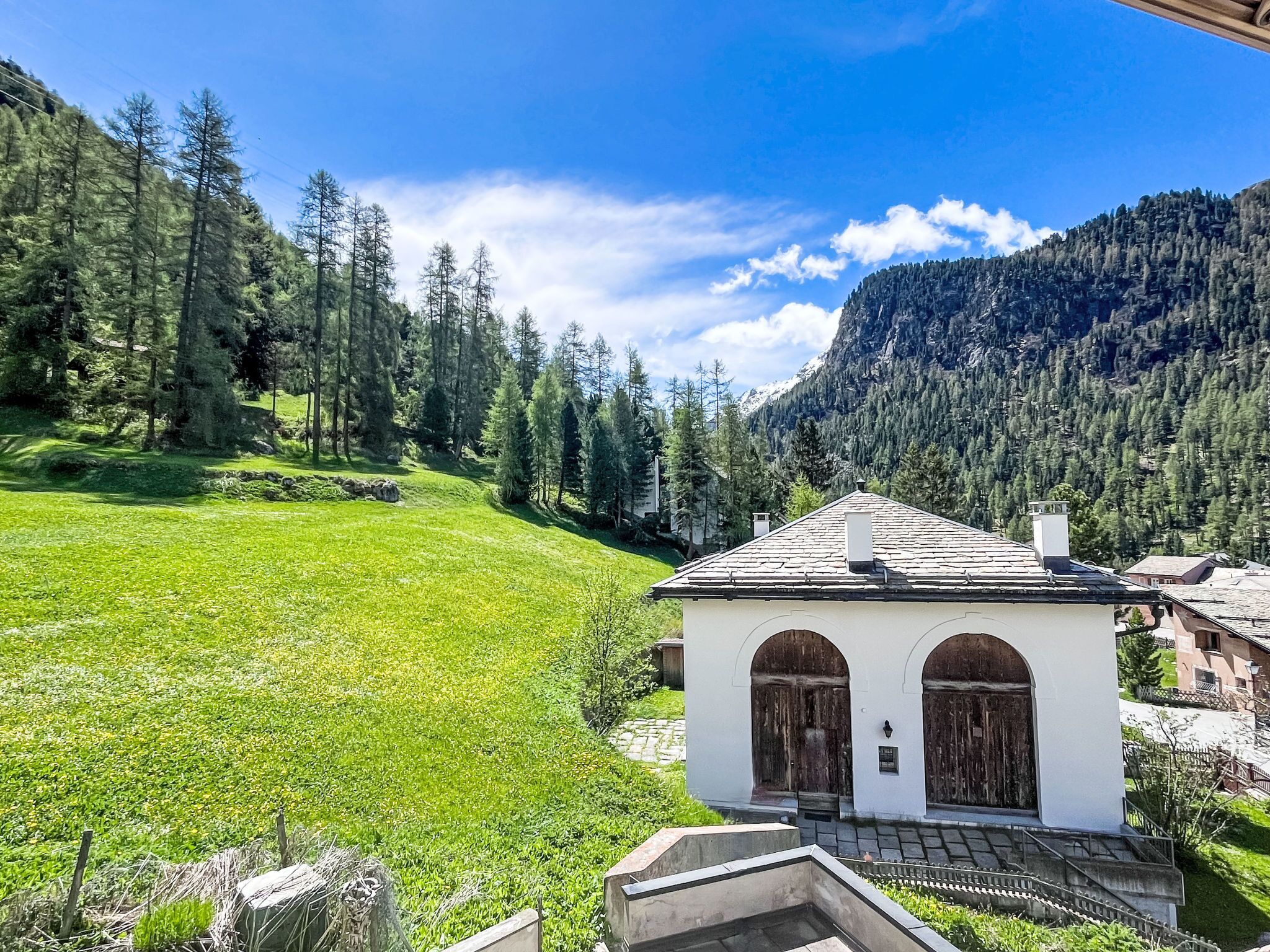 A white building with two arched doors, surrounded by a lush green field and a forested mountain backdrop.