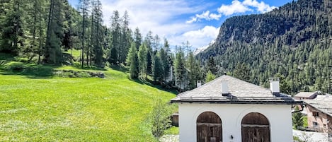 A white building with two arched doors, surrounded by a lush green field and a forested mountain backdrop.