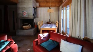 Interior acogedor de una cabaña con techo de madera, chimenea, mesa de comedor con sillas, sofá de cuero rojo y ventanas amplias con cortinas.