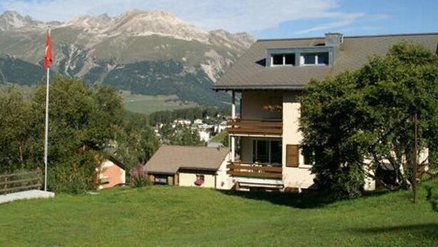 Une maison à deux étages dotée d’un balcon, entourée d’une pelouse verdoyante et d’arbres, avec des montagnes en arrière-plan.