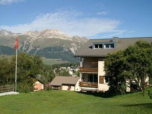 Une maison à deux étages dotée d’un balcon, entourée d’une pelouse verdoyante et d’arbres, avec des montagnes en arrière-plan.