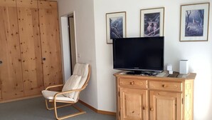 Ein Zimmer mit einem Holzschrank, einem gepolsterten Sessel, einem Fernseher auf einer Holzkommode und drei gerahmten Bildern an der Wand.