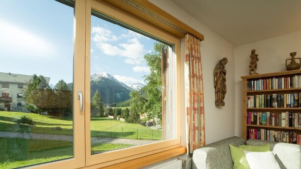 Ein Zimmer mit großem Fenster, einer Couch, einem Bücherregal und Blick auf einen Berg.
