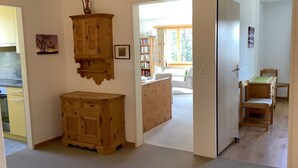 Ein Zimmer mit einem Holzkasten, einem kleinen Tisch mit Stühlen und einem Durchgang zu einem weiteren Raum mit Fenster und Bücherregal.