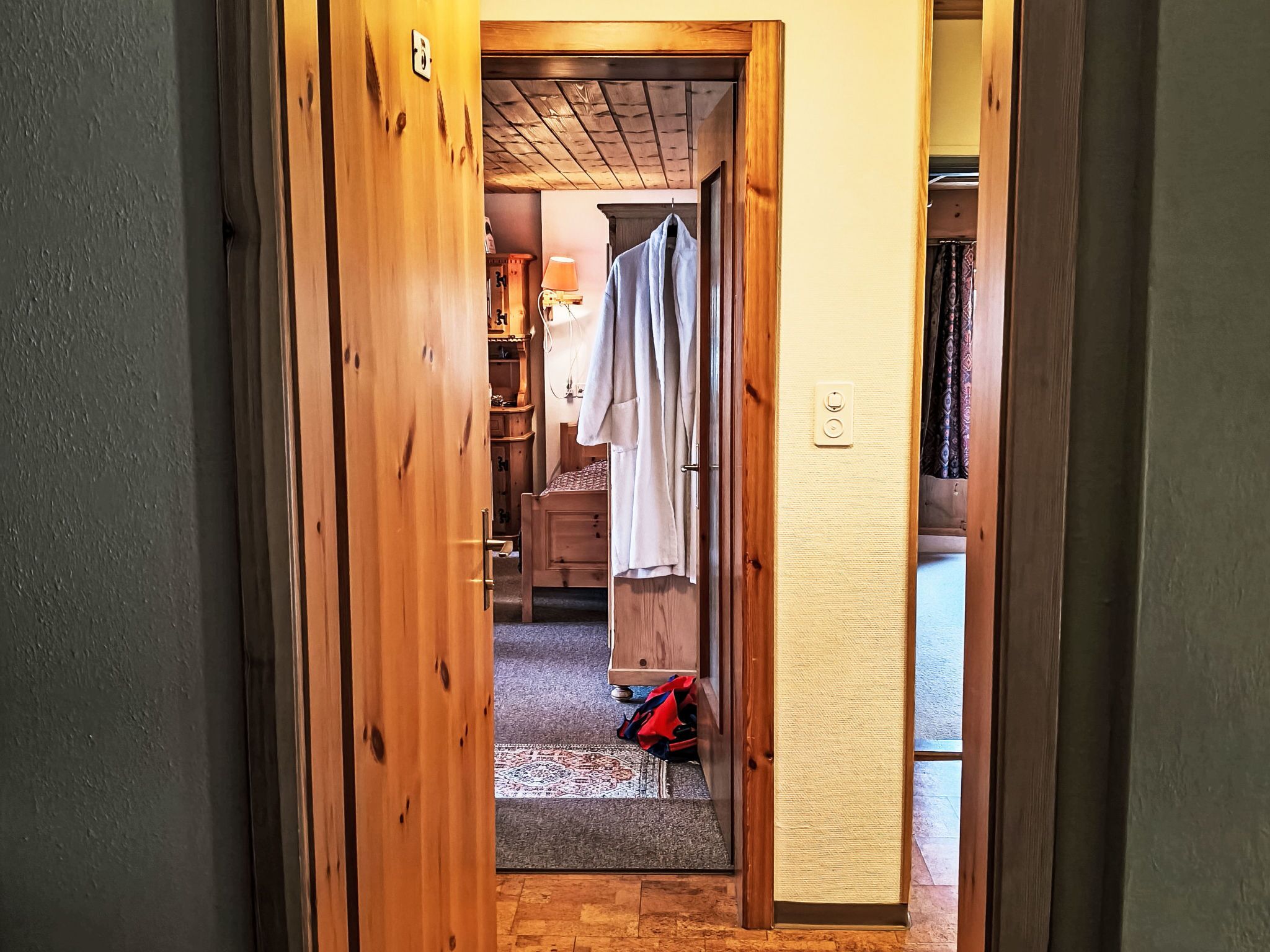 A wooden door leading to a room with a bed, a wardrobe, and a hanging garment.