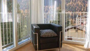 En balkon med en sort stol og et mønstret sædehynde, store vinduer der giver udsigt over et skovklædt bjerg.