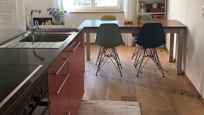 Et moderne køkken med en central ø, et spiseområde med to stole, en bogreol og et vindue med naturligt lys.