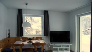 Uma sala de jantar com uma mesa e cadeiras de madeira, uma televisão sobre um suporte branco e uma janela com cortinas.