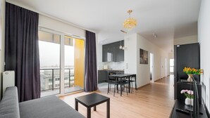 A modern living room with a sofa, a small table, and a dining area with chairs. There is a kitchen in the background with a dining table and chairs. The room has large windows with curtains and a chandelier. - Ursus Spacious Apartment (Warszawa)