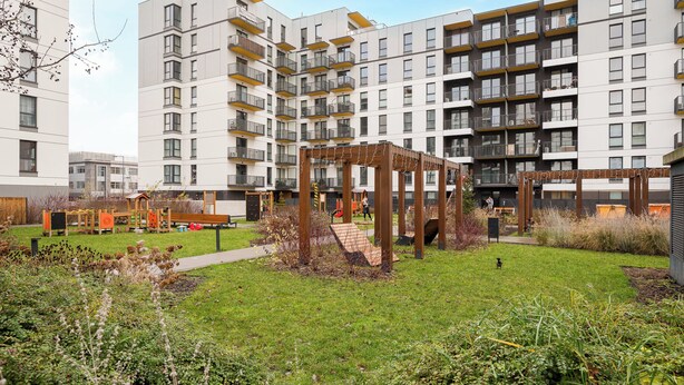A modern apartment complex with multiple floors, balconies, and a central courtyard with a wooden pergola and playground equipment. - Ursus Spacious Apartment (Warszawa)
