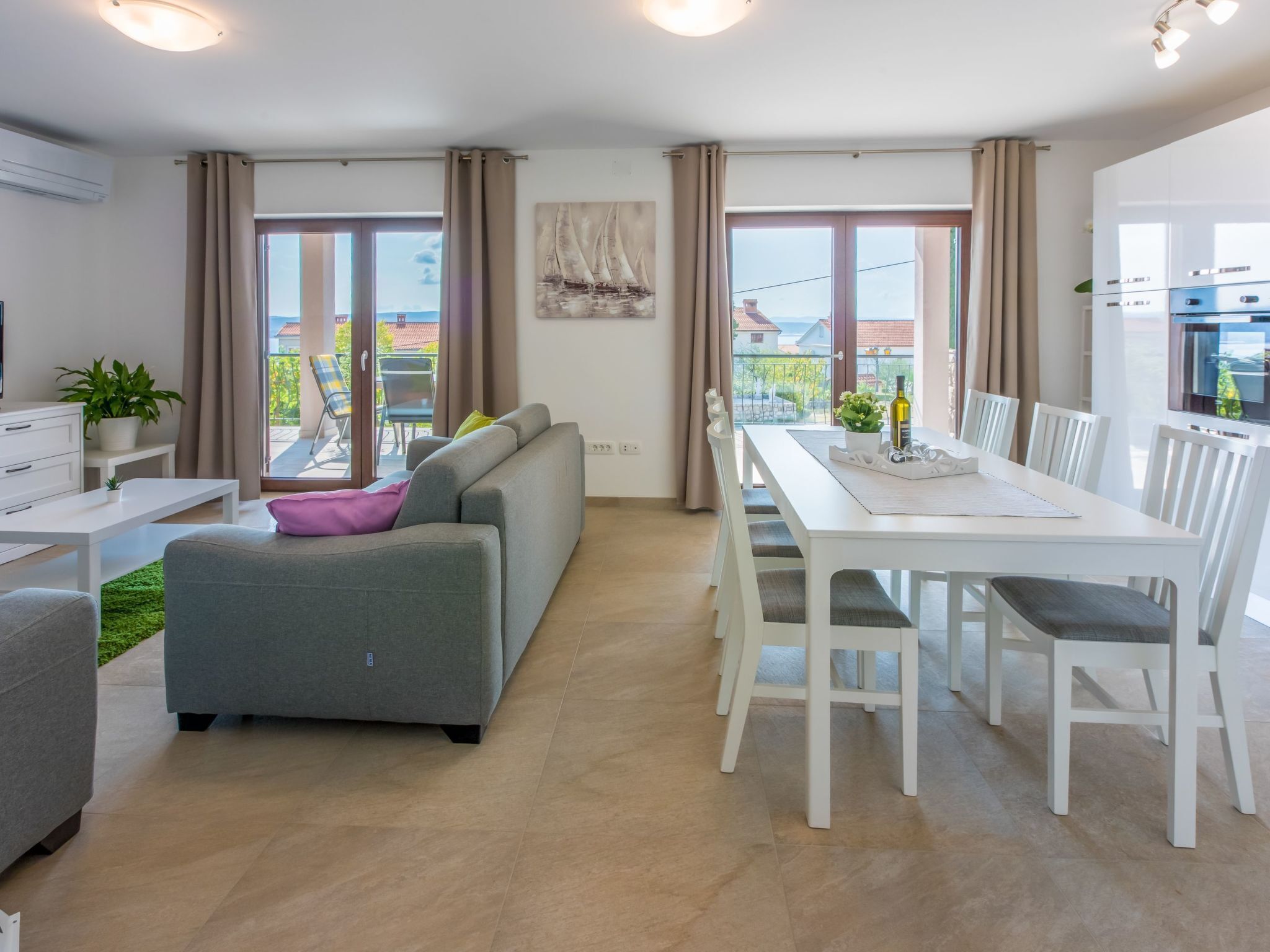 Un salon moderne avec un canapé gris, une table à manger avec des chaises blanches et une grande fenêtre avec des rideaux.