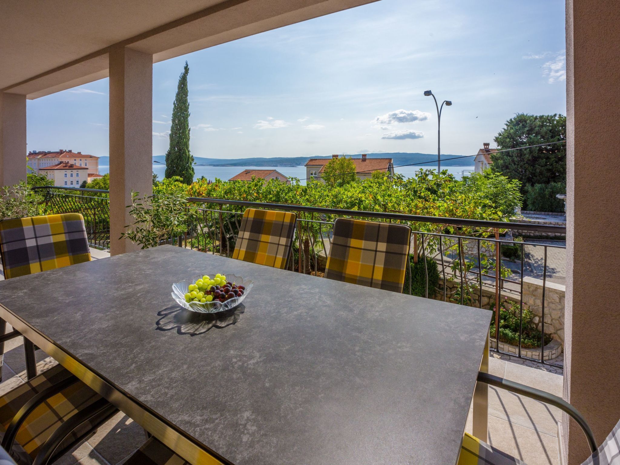 En balkon med et bord og stole, en skål med frugt og udsigt til havet og bygninger.