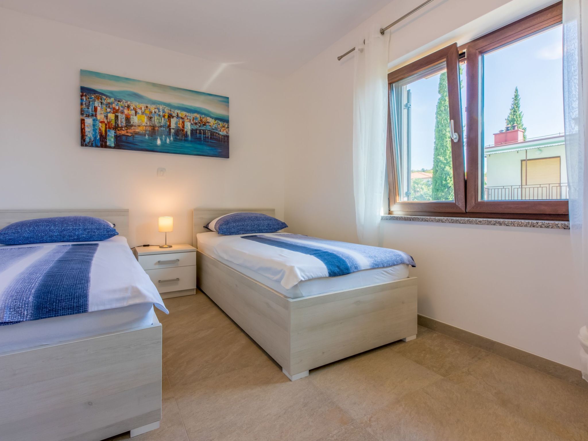 A bedroom with two beds, a nightstand, a painting, and a window with a view.