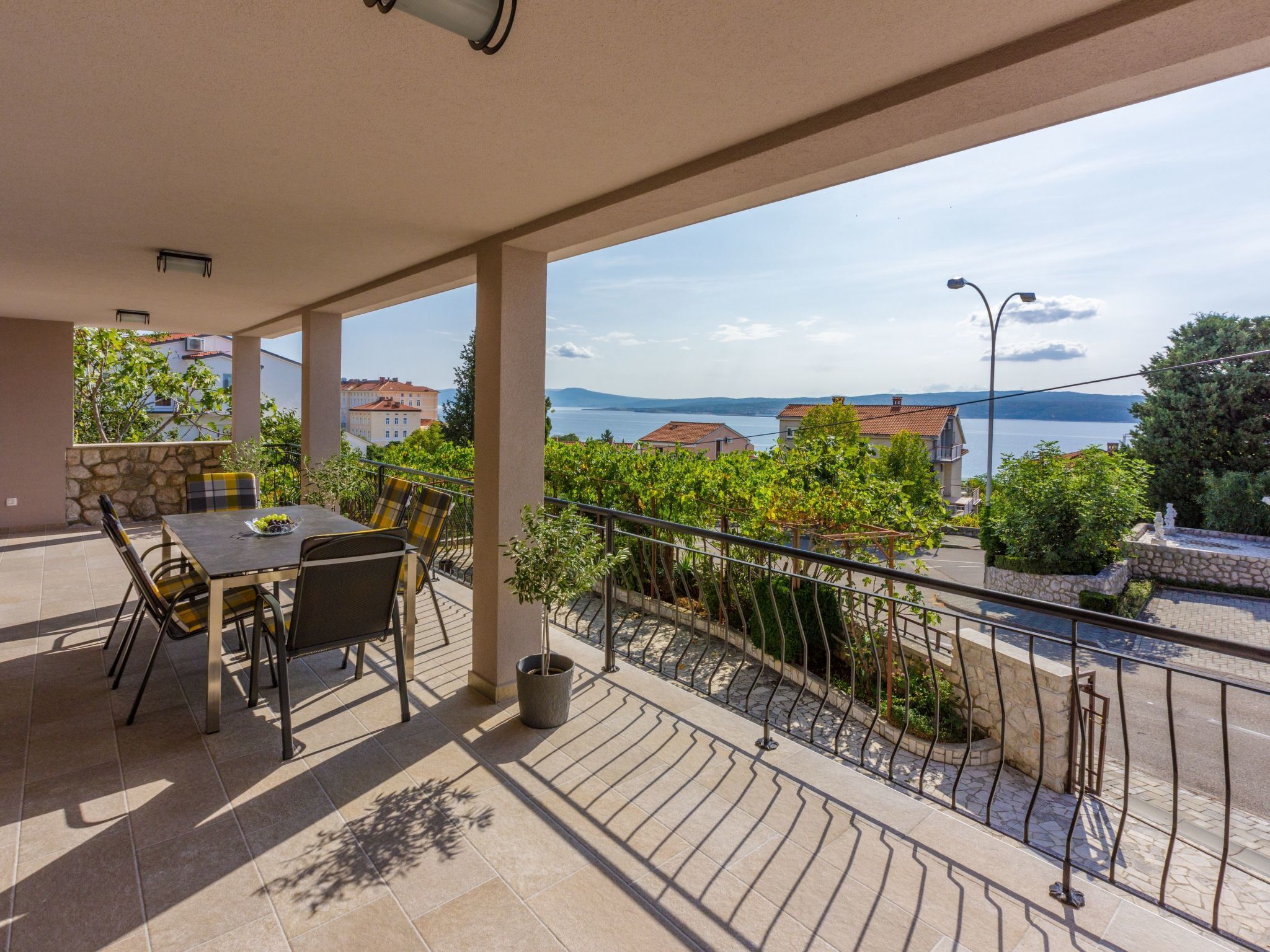 A covered outdoor area with a table and chairs, a stone wall, and a view of a body of water and buildings in the distance.